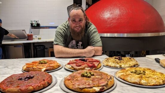 This photo shows Ferdinand Greco leaning against a table arrayed with various Argentine-style pizzas.