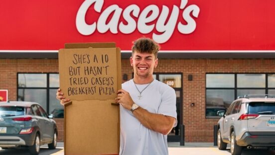 This photo shows a man standing in front of a Casey's store. He's holding up an empty pizza box with the following words written on the cardboard: "She's a 10 but hasn't tried Casey's breakfast pizza."
