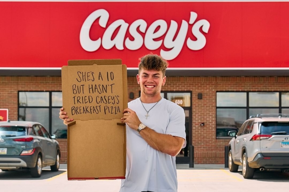 This photo shows a man standing in front of a Casey's store. He's holding up an empty pizza box with the following words written on the cardboard: "She's a 10 but hasn't tried Casey's breakfast pizza."
