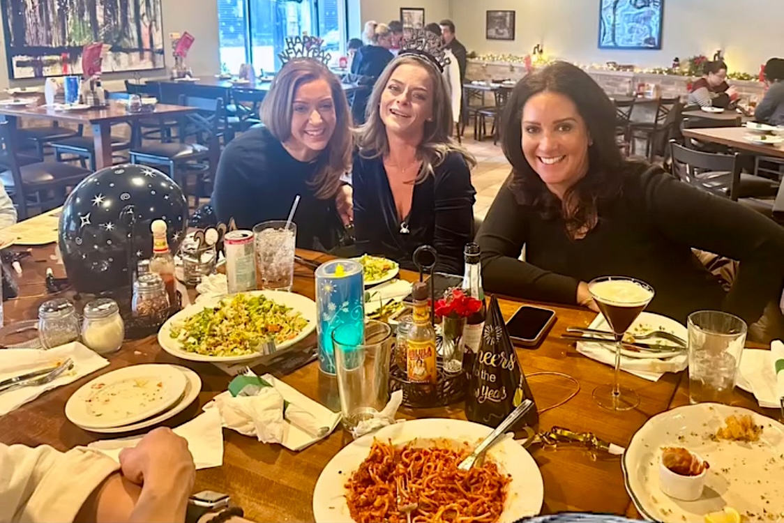 This photo shows three smiling women sitting at a table at Leona's with various dishes in front of them, including a big plate of spaghetti.