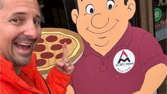 This close-up photo shows Matt Plapp in his trademark orange jacket posing with and pointing to a wooden statue of Aldo, owner of Aldo and Manny, in front of the pizzeria.