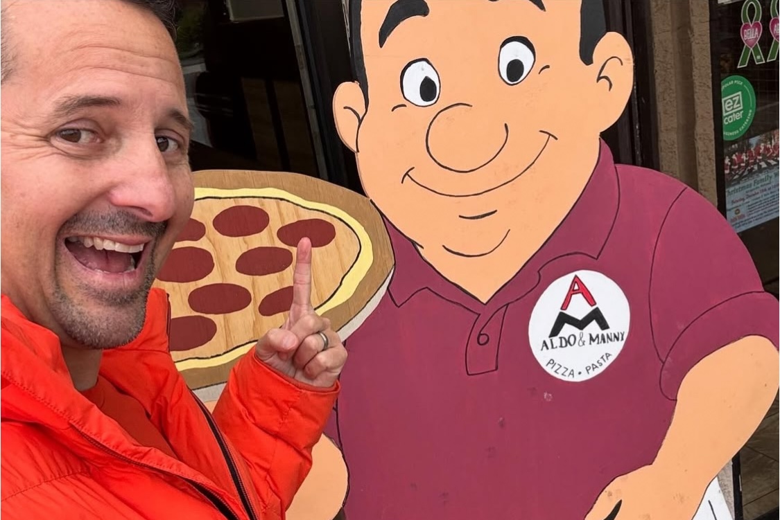 This close-up photo shows Matt Plapp in his trademark orange jacket posing with and pointing to a wooden statue of Aldo, owner of Aldo and Manny, in front of the pizzeria.