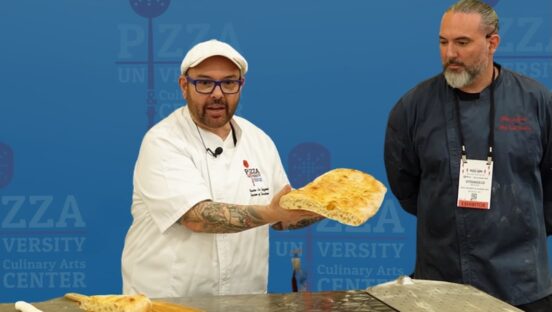 This photo shows Leo Spizzirri, in a white chef coat, holding a large piece of baked flatbread while Vitangelo Recchia, clad in a black chef coat, looks on.