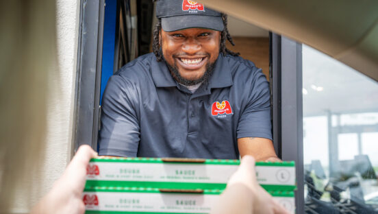 This photo shows a young male Marco's Pizza employee handing a customer a pizza through the drive-thru window.