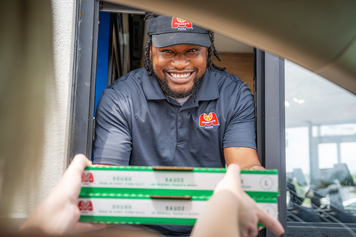 This photo shows a young male Marco's Pizza employee handing a customer a pizza through the drive-thru window.