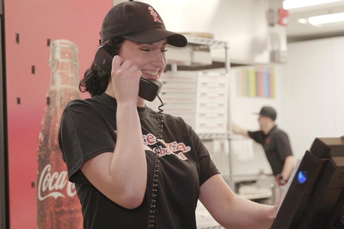 This photo shows a woman in a Master Pizza t-shirt smiling as she talks on the phone with a customer in one of the company's locations.