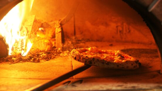 This photo shows a beautiful Neapolitan Pizza being turned around in a blazing wood-fired oven.