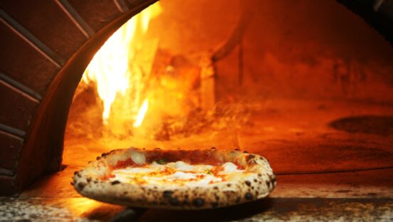 This photo shows a beautiful Neapolitan pizza perched on the edge of a flaming wood-fired oven.