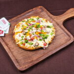 This photo shows a Paneer pizza with vegetables and cheese served on a wooden pizza dish.