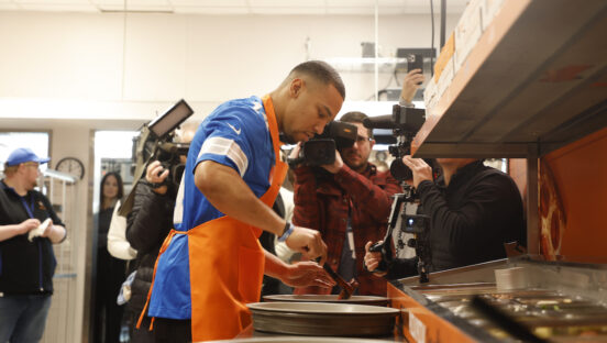 This photo shows St. Brown dipping a ladle into a vat of sauce at Little Caesars' headquarters while several photographers bunch around him and snap pictures.