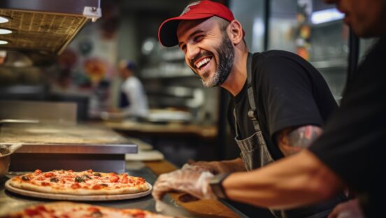 This photo shows a smiling male chef making pizza in the kitchen of a restaurant.