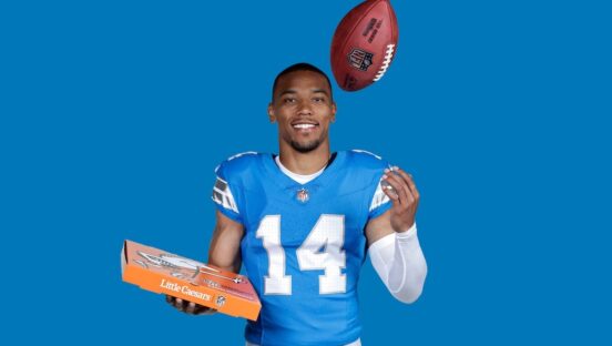 This photo shows Amon-Ra St. Brown in a blue No. 14 jersey, tossing a football in the air with one hand and holding a pizza box with the other.
