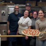 This photo shows a group of Alley Cat Pizzeria employees standing together in the kitchen, with one woman holding up a pepperoni pizza that's ready to go into the oven.