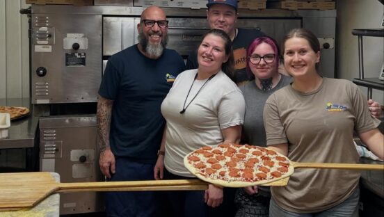 This photo shows a group of Alley Cat Pizzeria employees standing together in the kitchen, with one woman holding up a pepperoni pizza that's ready to go into the oven.