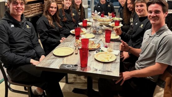 This photo shows a group of male and female athletes in black uniforms sitting together at a table.