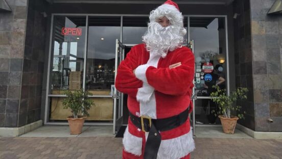 This photo shows Tore Trupiano, dressed as Santa Claus, standing in front of his restaurant with his arms crossed in a power pose.