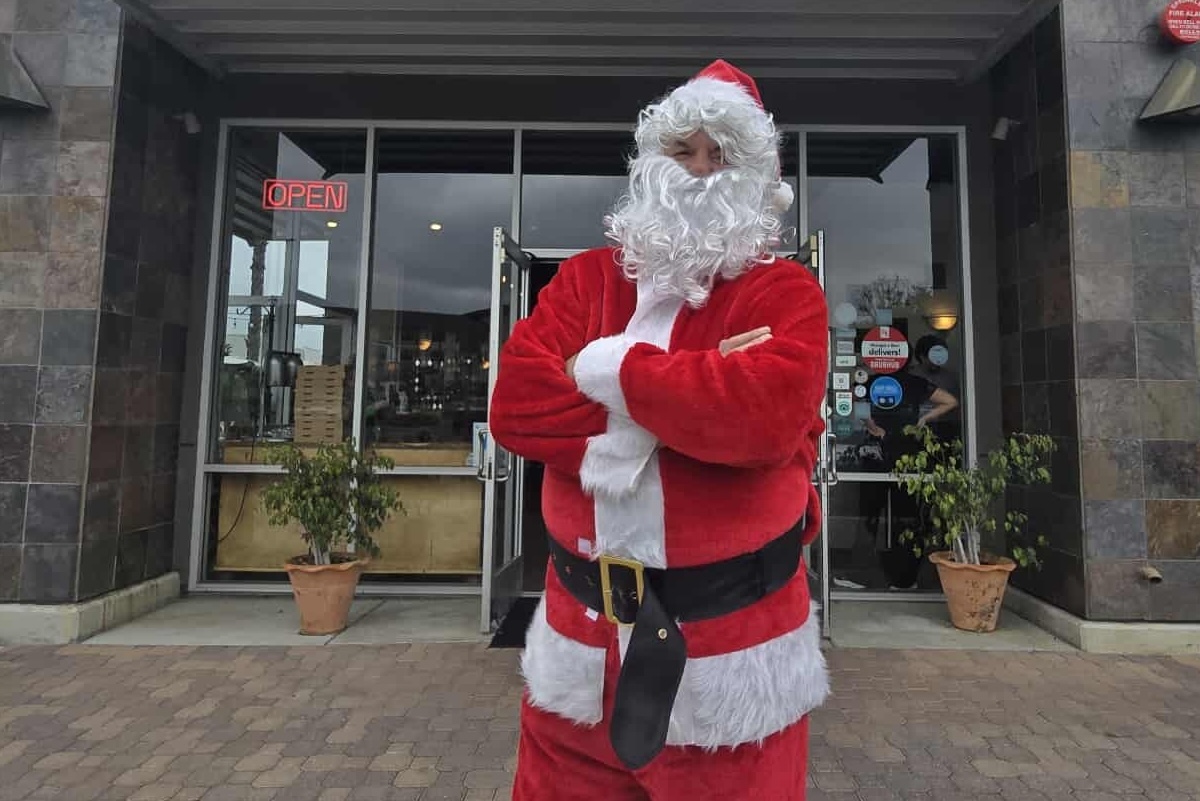 This photo shows Tore Trupiano, dressed as Santa Claus, standing in front of his restaurant with his arms crossed in a power pose.