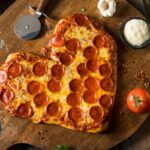 This photo shows a beautiful heart-shaped pizza topped with pepperoni. It's flanked by a tomato, a little serving dish of Parmesan and a bulb of garlic.
