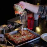 This photo shows a person's hand pulling out a slice of pizza topped with pepperoni and jalapeños, with a nice cheese pull.