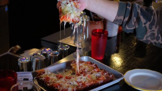 This photo shows a person's hand pulling out a slice of pizza topped with pepperoni and jalapeños, with a nice cheese pull.