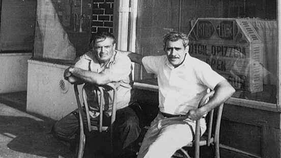 This vintage black and white photo shows two middle-aged men seated in chairs outside Totonno's, perhaps in the 1950s.