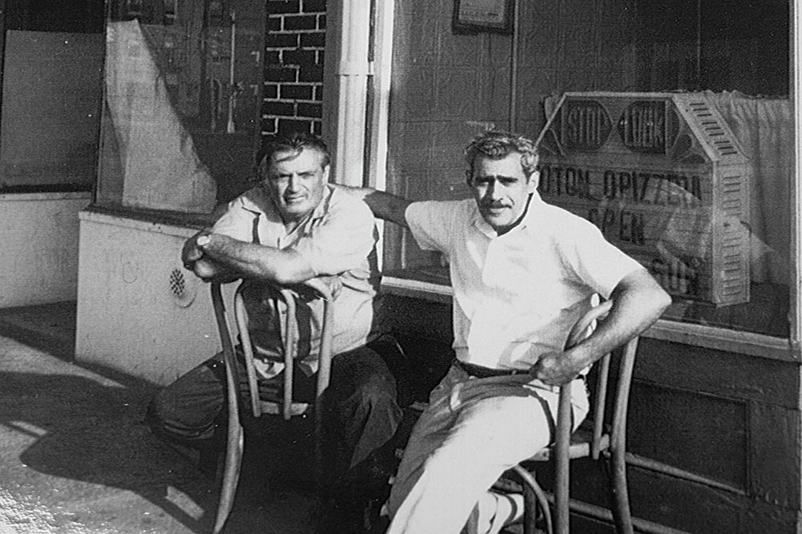 This vintage black and white photo shows two middle-aged men seated in chairs outside Totonno's, perhaps in the 1950s.
