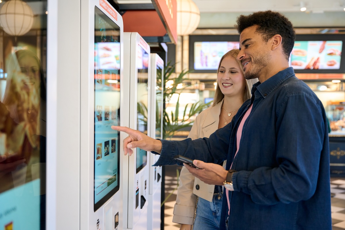 This photo shows a young couple smiling and selecting food on a modern self service touch screen kiosk in a casual fast food restaurant setting.