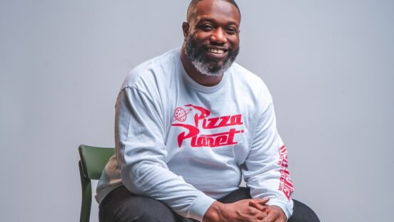 This photo shows Tyrell Reed wearing a Pizza Planet sweater and sitting in a chair with a big smile.