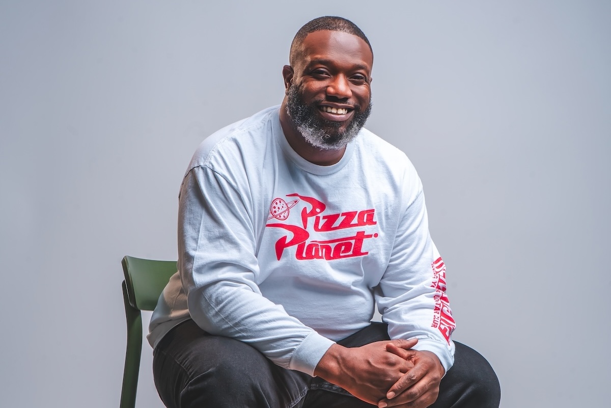 This photo shows Tyrell Reed wearing a Pizza Planet sweater and sitting in a chair with a big smile.