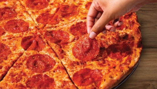 This photo shows a woman's finger picking a slice of pepperoni off a whole pepperoni pizza.