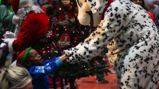 This photo shows a person in a Dalmatian-themed mascot costume holding hands with a happy-looking girl wearing a Santa hat at a holiday party.
