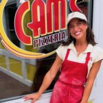This photo shows Giuliana in a white shirt, red apron and white visor standing in front of the Cam's Pizzeria sign at one of the brand's locations.