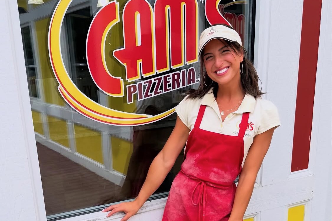 This photo shows Giuliana in a white shirt, red apron and white visor standing in front of the Cam's Pizzeria sign at one of the brand's locations.