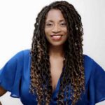 This photo shows Benita Gibson in a blue blouse, hands on her hips and smiling for the camera.