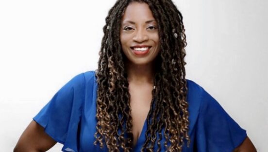 This photo shows Benita Gibson in a blue blouse, hands on her hips and smiling for the camera.