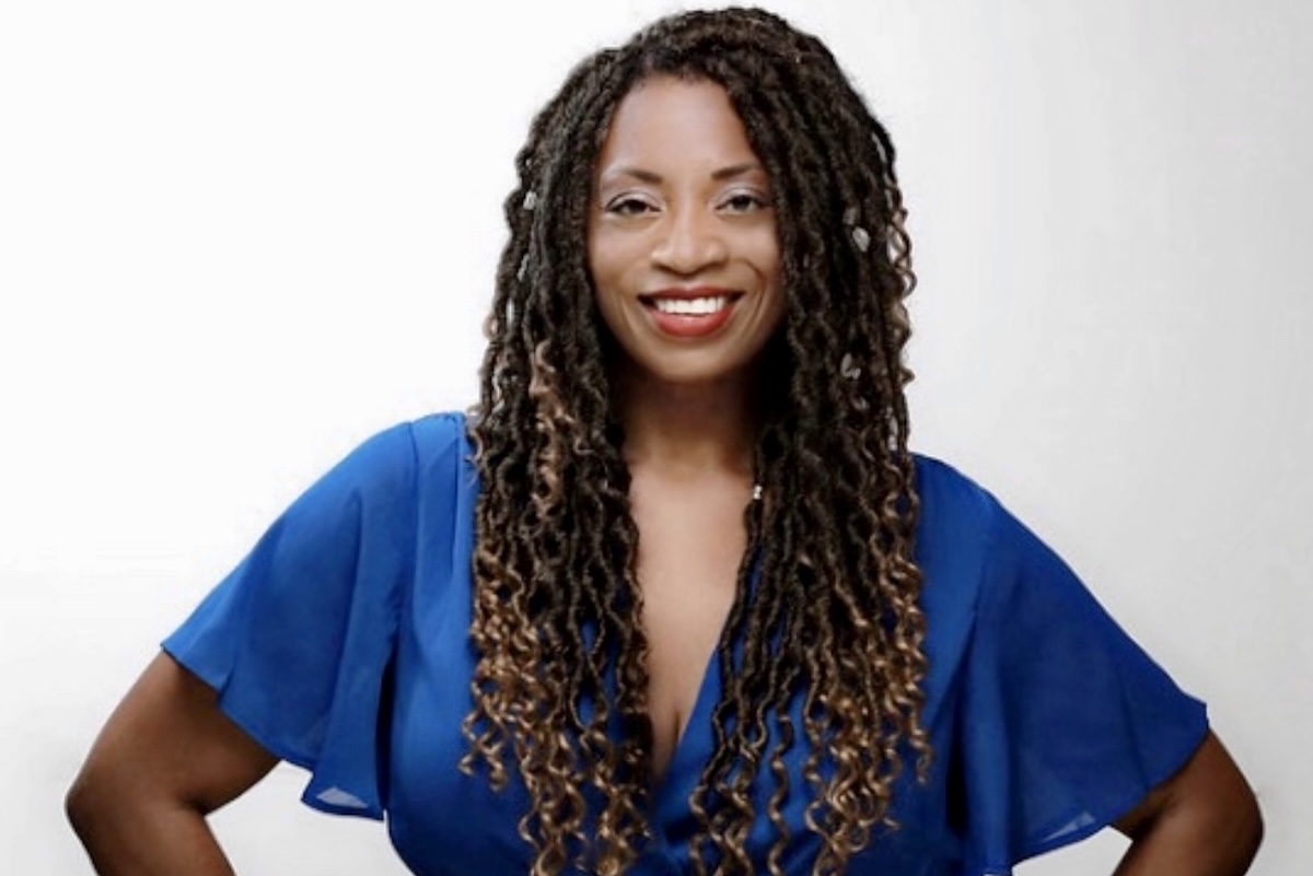 This photo shows Benita Gibson in a blue blouse, hands on her hips and smiling for the camera.