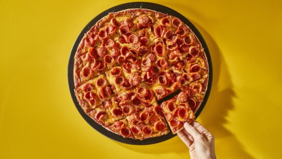 This photo shows a St. Louis-style pepperoni pizza against a dark yellow background. A woman's hand is removing a slice from the whole pie.