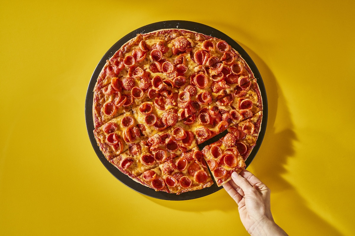 This photo shows a St. Louis-style pepperoni pizza against a dark yellow background. A woman's hand is removing a slice from the whole pie.