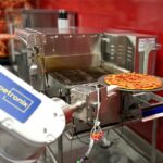 This photo shows Pepptron's robotic arm removing a baked pepperoni pizza from the conveyor oven.