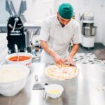 This photo shows a man topping pizzas at a prep table in a restaurant.
