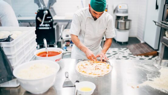 This photo shows a man topping pizzas at a prep table in a restaurant.