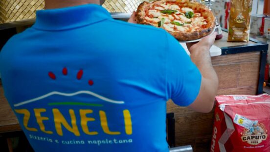 This photo shows a man in a blue Zeneli shirt, seen from behind and holding a Margherita pizza.