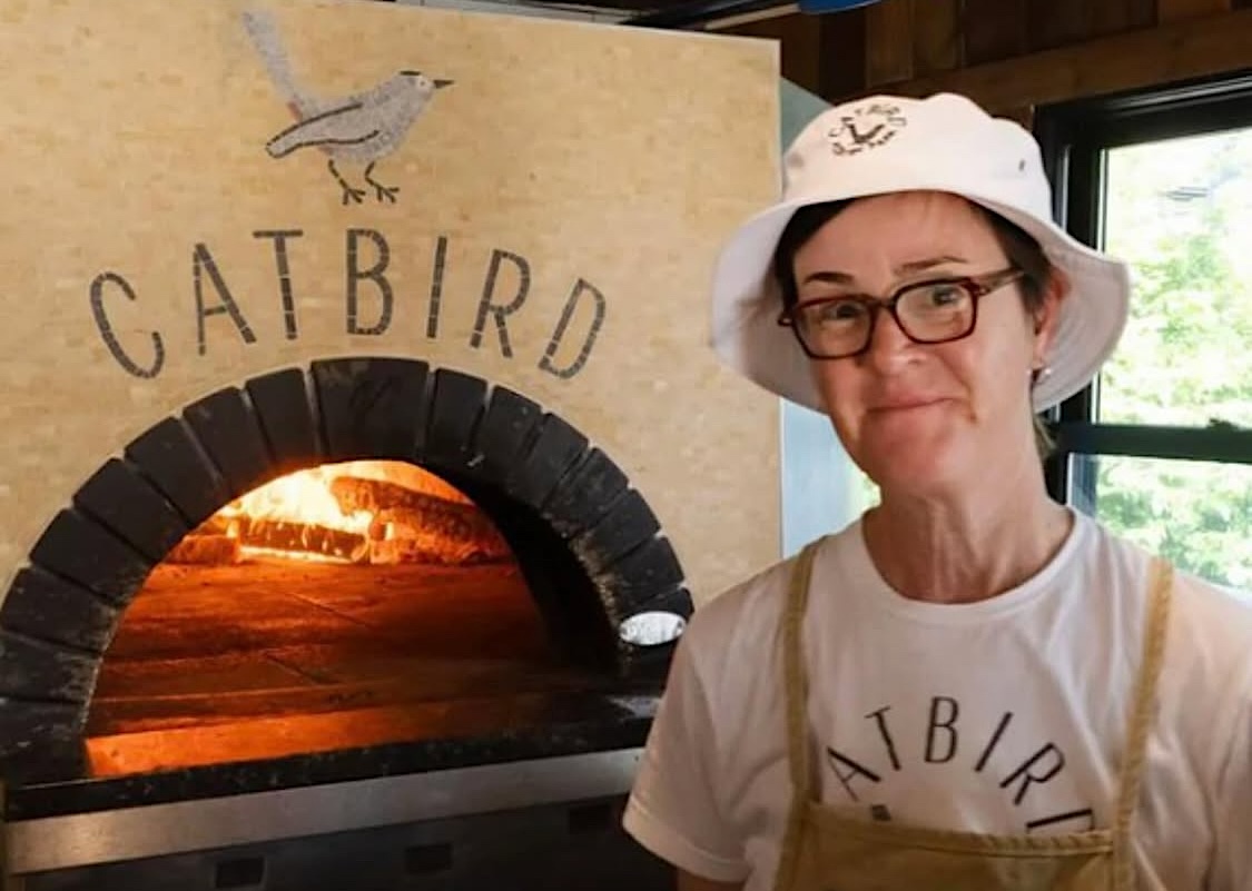 This photo shows a grinning Aimee McElroy standing in front of of the wood-fired oven which has the Catbird logo on the front.