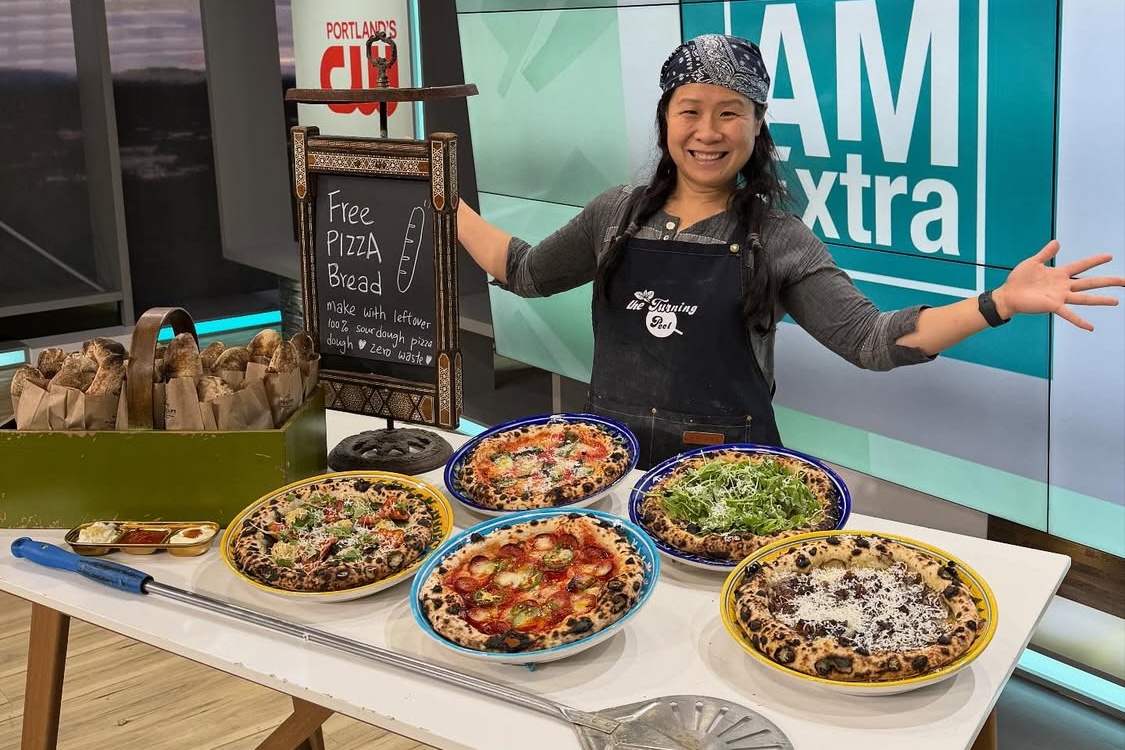 This photo shows Candy Yiu, smiling and her arms spread wide in a welcoming gesture, on the set of a local TV news show, with five pizzas laid out on a table in front of her.