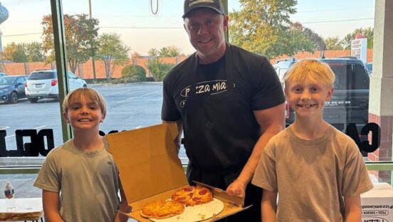 This photo shows Dave Dombrowski holding up a box of pizza with two young blonde-haired boys beside him.
