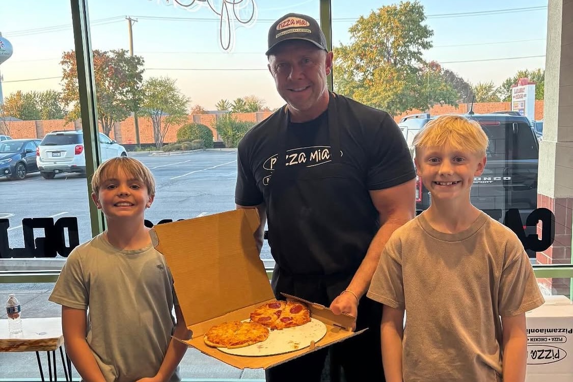 This photo shows Dave Dombrowski holding up a box of pizza with two young blonde-haired boys beside him.