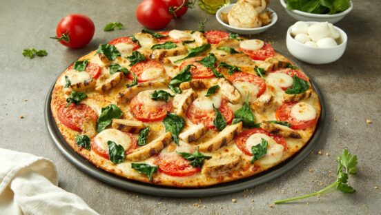 This photo shows a pizza topped with chicken, spinach, roasted garlic, tomato slices and mozzarella.