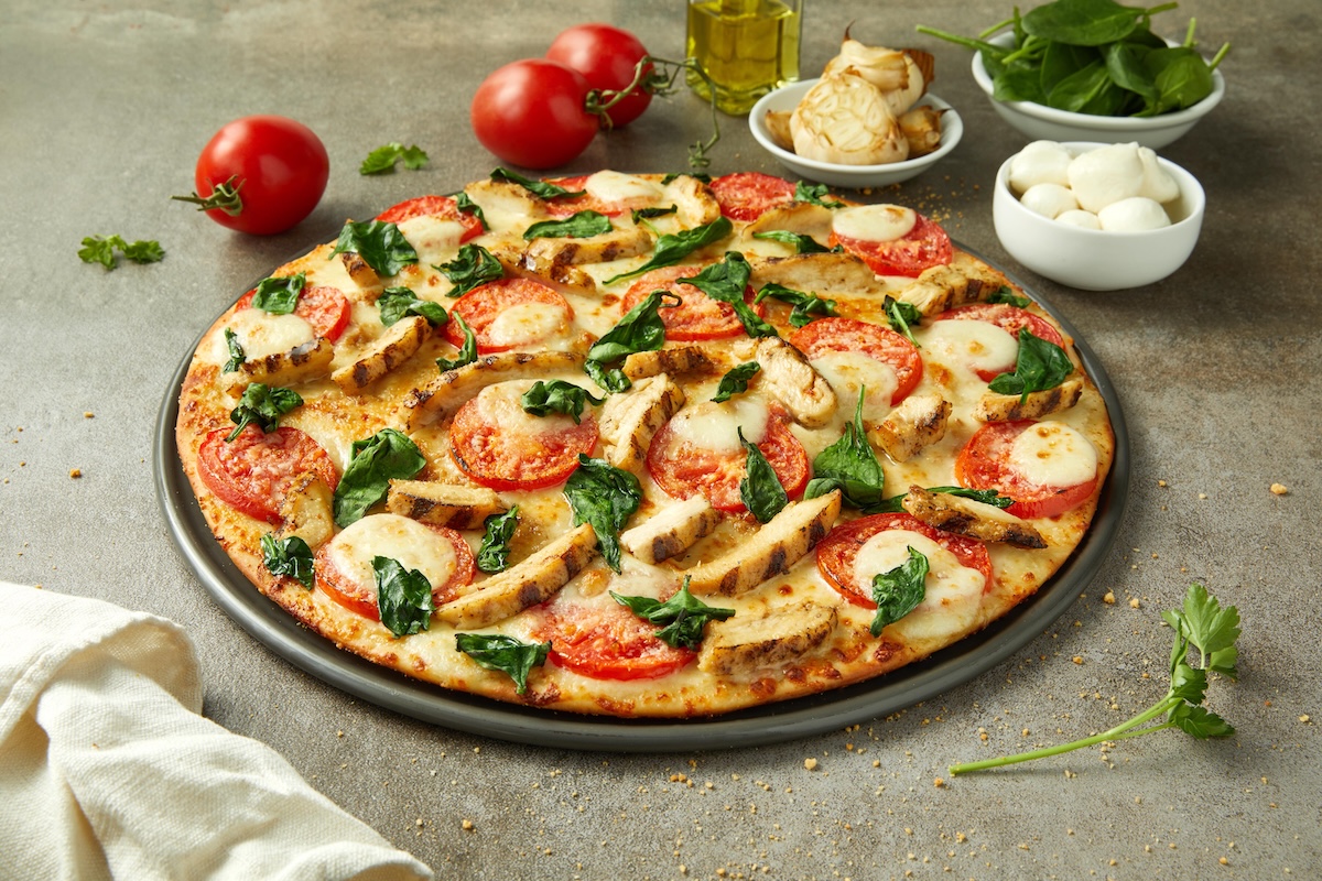 This photo shows a pizza topped with chicken, spinach, roasted garlic, tomato slices and mozzarella.