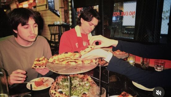 This photo shows two young men with dark hair eating pizzas from a double-stacked tray of pies at See No Evil Pizza.
