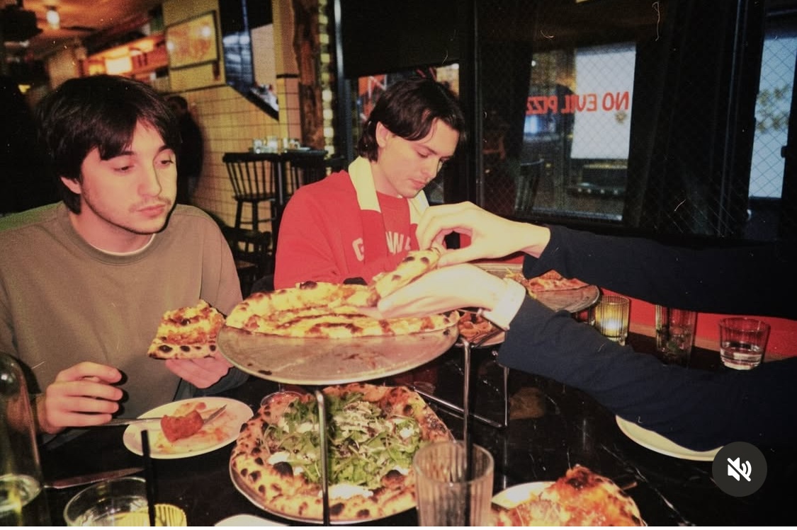 This photo shows two young men with dark hair eating pizzas from a double-stacked tray of pies at See No Evil Pizza.
